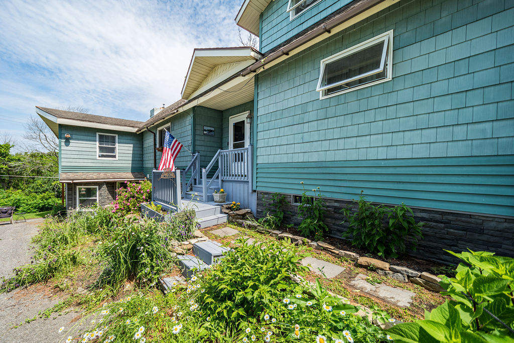 Front exterior of the teal-shingled home with flag and garden beds